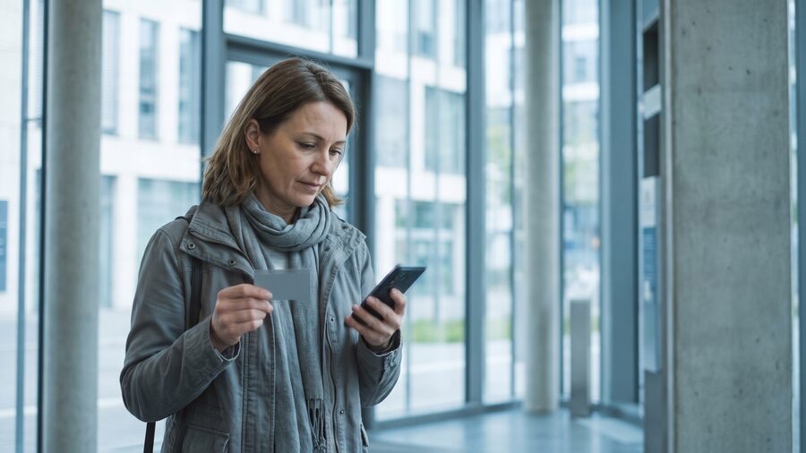 Frau pr&uuml;ft ihre Bankkarte unterwegs mit dem Smartphone in der Hand
