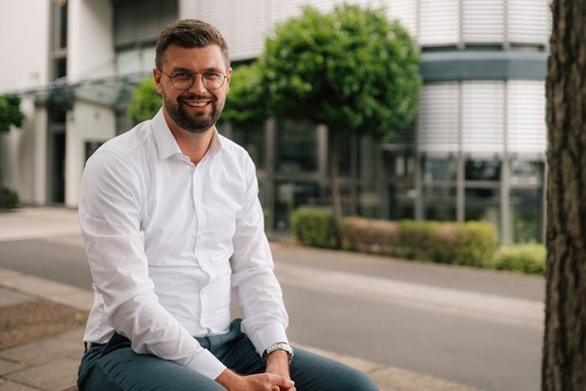 Portr&auml;t eines l&auml;chelnden Mannes mit Brille vor einem B&uuml;rogeb&auml;ude