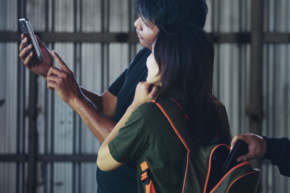 Zwei Männer lenken eine Frau mit einem Smartphone ab, während eine dritte Hand in ihren Rucksack greift