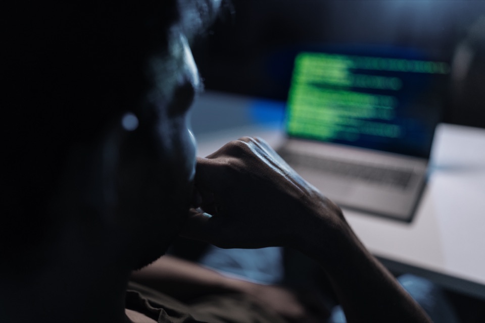 Person sitzt im Dunkeln vor einem Laptop, auf dessen Bildschirm grüner Programmcode leuchtet