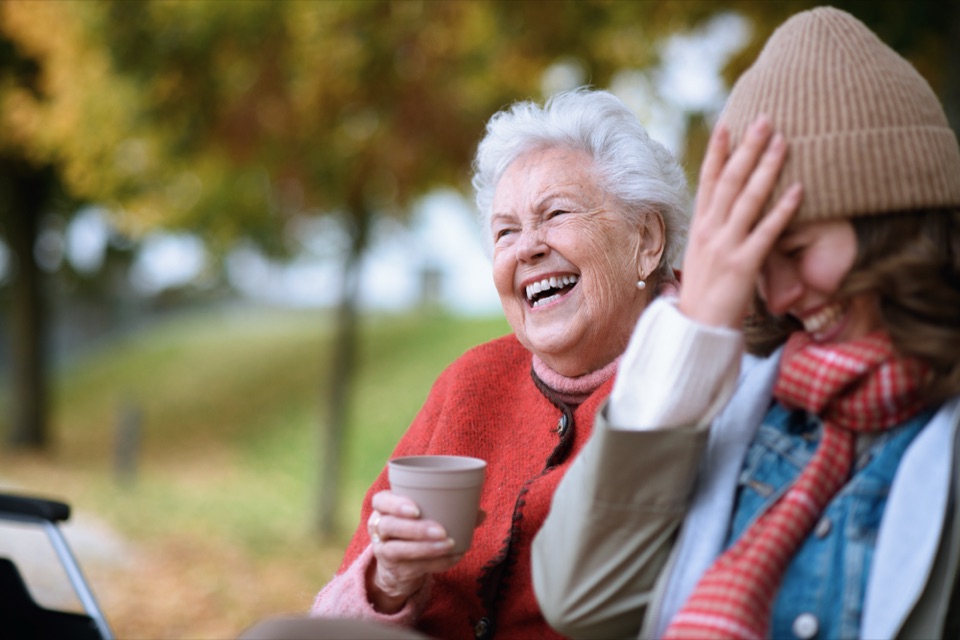 Lachende Seniorin mit einer Tasse in der Hand sitzt neben einer jungen Frau im herbstlichen Park