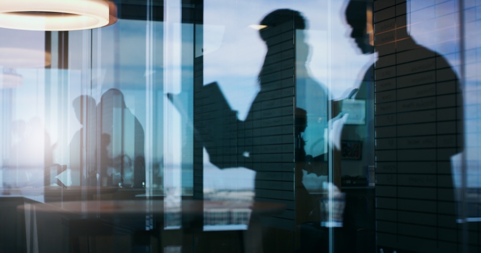 Silhouetten mehrerer Menschen hinter einer verspiegelten Glasfassade in einem Büro